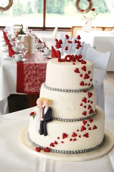 a wedding cake with red hearts on it and a bride and groom sitting on top