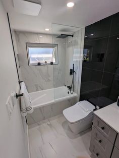 a white toilet sitting next to a bath tub in a bathroom under a window on top of a tiled floor