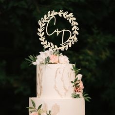 a three tiered cake with flowers and leaves on the top is decorated with an acrylic monogram