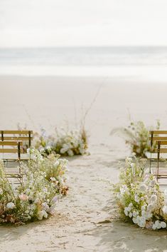 two wooden benches sitting next to each other on a sandy beach near the ocean with flowers