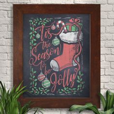 a chalkboard christmas stocking sign hanging on a brick wall next to a potted plant