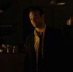 a man wearing a suit and tie standing in a dark room with bottles on the counter