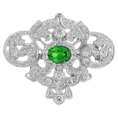 This is an impressive vintage inspired brooch pendant set in 18k white gold featuring a gorgeous oval cut green tsavorite gemstone measuring 8 x 6 mm. The brooch is further set with stunning round cut diamonds of H color and SI clarity. Super sparkly! Brooch Information Metal: 18K White Gold Width: 40 mm. Length: 33 mm. Weight: 14.10 g. (approx. in total) Center Gemstones Type: Tsavorite Shape: Oval Size: 8 x 6 mm. Number: 1 Weight: 1.50 Carat (approx.) Accent Gemstones Type: Diamond Shape: Roun Elegant Green Diamond Brooch, Classic Diamond Brooch With Gemstones, Classic Diamond Gemstone Brooches, Heirloom Oval Gemstone Brooches, Elegant Oval Tsavorite Jewelry, Elegant Oval Gemstone Brooches, Elegant Green Gemstone Brooch, Elegant Green Gemstone Brooches, Elegant Green Oval Brooches