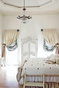 a white bed sitting under a chandelier in a bedroom next to two windows