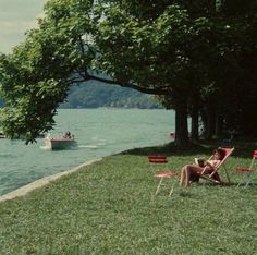 two people sitting in lawn chairs on the grass next to water with boats and trees