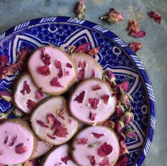 a blue and white plate topped with cookies covered in pink frosting next to dried rose petals