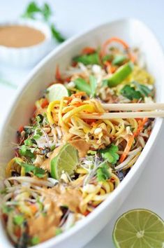 a white dish filled with noodles, vegetables and lime wedges next to some dipping sauce