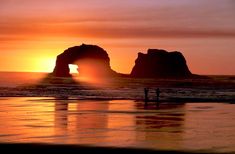 the sun is setting behind two large rock formations on the beach with people standing in the water