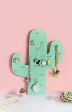 a green cactus shaped wall hanging on a pink wall next to other jewelry and bracelets