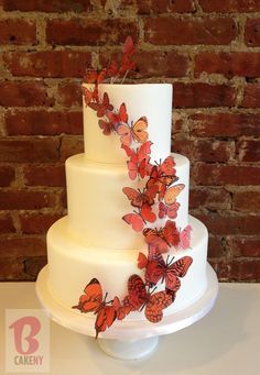 a three tiered cake with butterflies on the icing and frosting is displayed in front of a brick wall