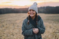 a woman standing in a field wearing a hat and scarf with her arms folded out