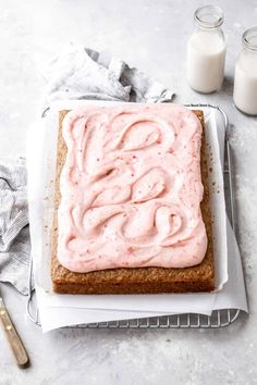 a square cake with pink frosting sitting on top of a cooling rack next to two glasses of milk