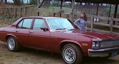 a woman standing next to a red car in the dirt near a fenced area