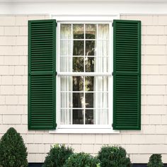 an open window with black shutters on a white brick building in front of green bushes