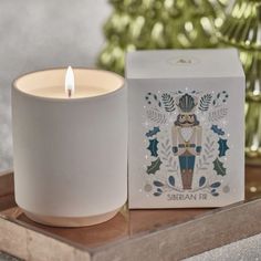 a white candle sitting on top of a wooden tray next to a box with a lit candle in it