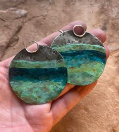 a pair of green and blue earrings sitting on top of a handmade piece of metal