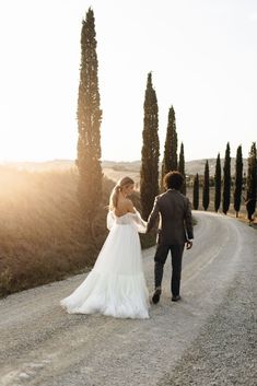 a bride and groom walking down the road in front of some tall trees at sunset