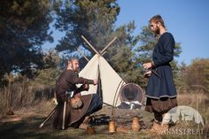 two men dressed in period clothing sitting around a campfire with an old fashioned teepee