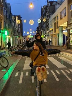 two people riding bikes down a street at night with christmas lights strung above the buildings