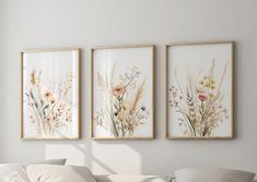 three floral paintings hang on the wall above a white couch in front of a window