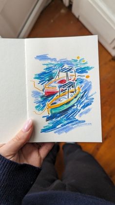 a person holding an open book with watercolors on it and a boat in the background