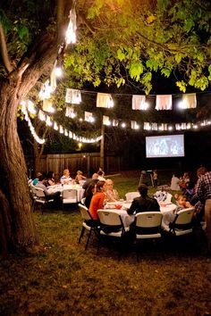 people are sitting at tables in the yard under lights and paper lanterns strung from trees