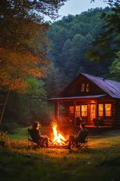 two people sitting around a campfire in front of a log cabin with the lights on