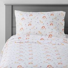 a bed with white sheets and colorful unicorns on the sheet set is in front of a gray headboard