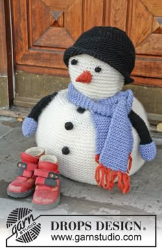 a crocheted snowman sitting on the ground next to a wooden door wearing a hat and scarf