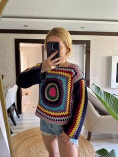 a woman taking a selfie in front of a mirror wearing shorts and a crocheted sweater