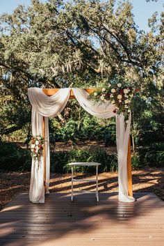 an outdoor wedding setup with white drapes and flowers on the arbor, surrounded by trees