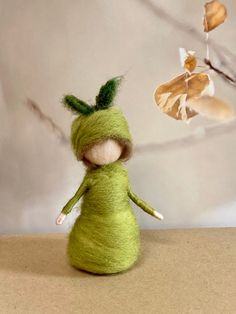 a small green doll sitting on top of a table next to a branch with leaves