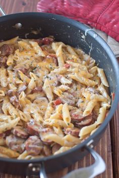 a skillet filled with pasta and sausage on top of a wooden table next to a red towel