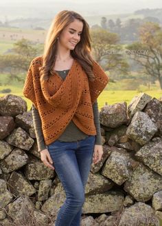 a woman standing next to a stone wall wearing a brown knitted sweater and jeans