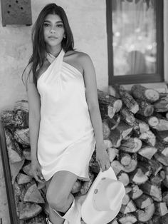 a woman in a white dress and cowboy hat leaning against a pile of firewood