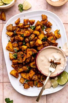 a white plate topped with fried potatoes next to a bowl of ranch dressing and lime wedges