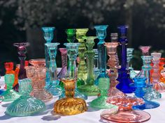 many different colored glass vases sitting on a white tablecloth with trees in the background