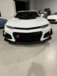 the front end of a white sports car parked in a garage with other cars behind it