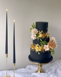 a black and gold wedding cake with flowers on it, next to two tall candles