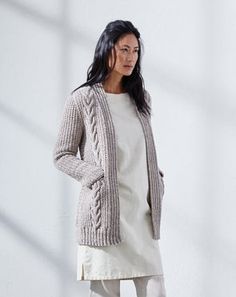 a woman standing in front of a white wall wearing a gray cardigan sweater and beige pants