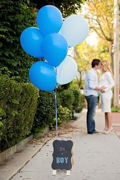 a couple kissing on the sidewalk with blue balloons attached to their feet and a sign that says it's a boy