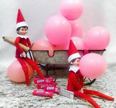 two elfs are sitting in front of balloons and a cart with pink balls on it