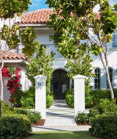 the front entrance to a white house with red flowers and trees on either side of it