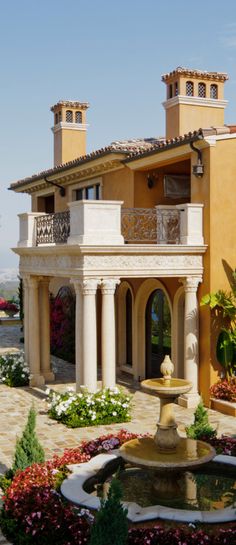 a house with a fountain in front of it and lots of flowers on the ground