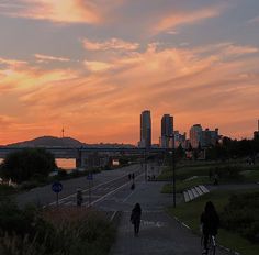 two people are walking down the sidewalk in front of some buildings and water at sunset