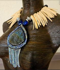 A labrodorite or spectrolite sculpted pendant with sapphire blue and silver aurora borealis rhinestone fringe is the focal point on this one-of-a-kind statement necklace or chest piece. Flanking the pendant are two sapphire blue crystals, silver aurora borealis rhinestone rondelles and stamped silver tone tribal bicones. The remainder of the necklace is blonde coconut fringe with black lava beads, silver tone metal bi-cones and blue crystals finishing the back. This lightweight and comfortable p Artisan Sapphire Jewelry, Bohemian Jewelry With Gemstone Accents And Labradorite, Unique Labradorite Necklace With Large Stone, Unique Labradorite Stone Necklaces, Artisan Labradorite Gemstone Bead Jewelry, Unique Labradorite Necklaces With Stones, Exotic Dramatic, Black Aurora, Jewellery Shop Design