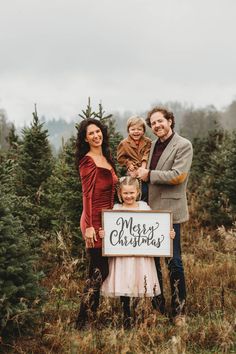 a family holding a merry christmas sign in the middle of a field with pine trees