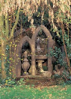 an old stone fountain in the middle of a garden surrounded by trees and bushes with flowers hanging from it