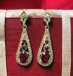 "Three rich merlot-colored garnets dangle from each of these push post dangle and drop earrings. Four are round-faceted garnets and two are larger oval shapes. The earring itself is a tear-shaped dangle that swings and sways with your every movement! These earrings are a bright shiny gold color with a rope motif outline.   The total length is 1 1/2\" Garnet is the birthstone for January. Spiritual Meaning of Garnet Garnet is also said to inspire contemplation and truthfulness and offers its wear Evening Ruby Dangle Jewelry, Elegant Garnet Drop Earrings, Elegant Garnet Earrings, Formal Ruby Earrings In Yellow Gold, Elegant Gold Garnet Earrings, Gold Ruby Earrings For Formal Occasions, Formal Gold Ruby Earrings, Formal Ruby Yellow Gold Earrings, Formal Yellow Gold Ruby Earrings