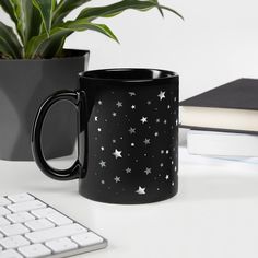 a black coffee mug with white stars on it next to a keyboard and potted plant